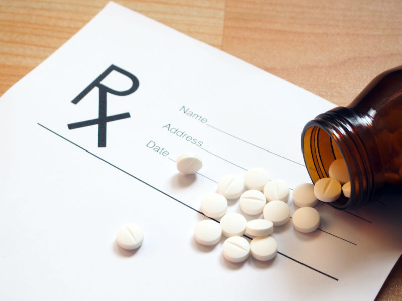 White tablets spilling out of brown glass bottle with blank Rx prescription form on wood table. Health care and medical concept. Selective focus.